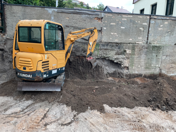 Gelbes Baufahrzeug gräbt Erde an einer Wand.